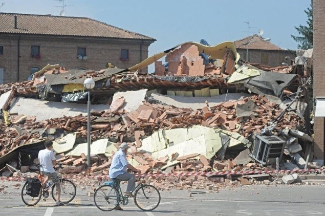 Dvojica cyklistov hľadí na zrútené domy a trosky, ktorých sú plné mestá