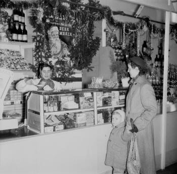 Zákazníčka nakupuje tovar vo vyzdobenej mestskej tržnici v Bratislave, ktorá ponúka rôzne dobroty.  (1955)