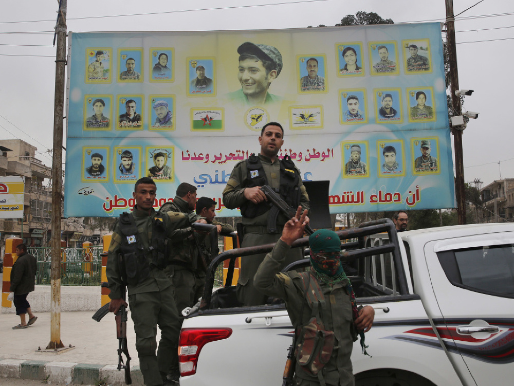 Fotografia z 28. marca 2018 členovia kurdských vnútorných bezpečnostných síl stoja na svojom vozidle pred obrovským plagátom zobrazujúcim portréty bojovníkov zabitých proti islamskej štátnej skupine v Manbídži na severe Sýrie.