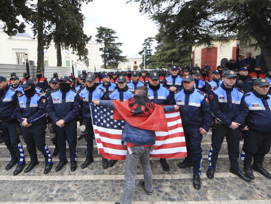 Protestujúci s albánskou zástavou na chrbte máva americkou zástavou pred albánskymi policajtmi 