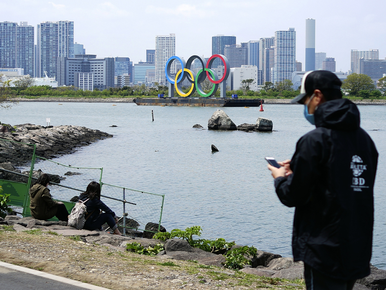 Na snímke ľudia s ochrannými rúškami oddychujú pred olympijskými kruhmi v Tokiu