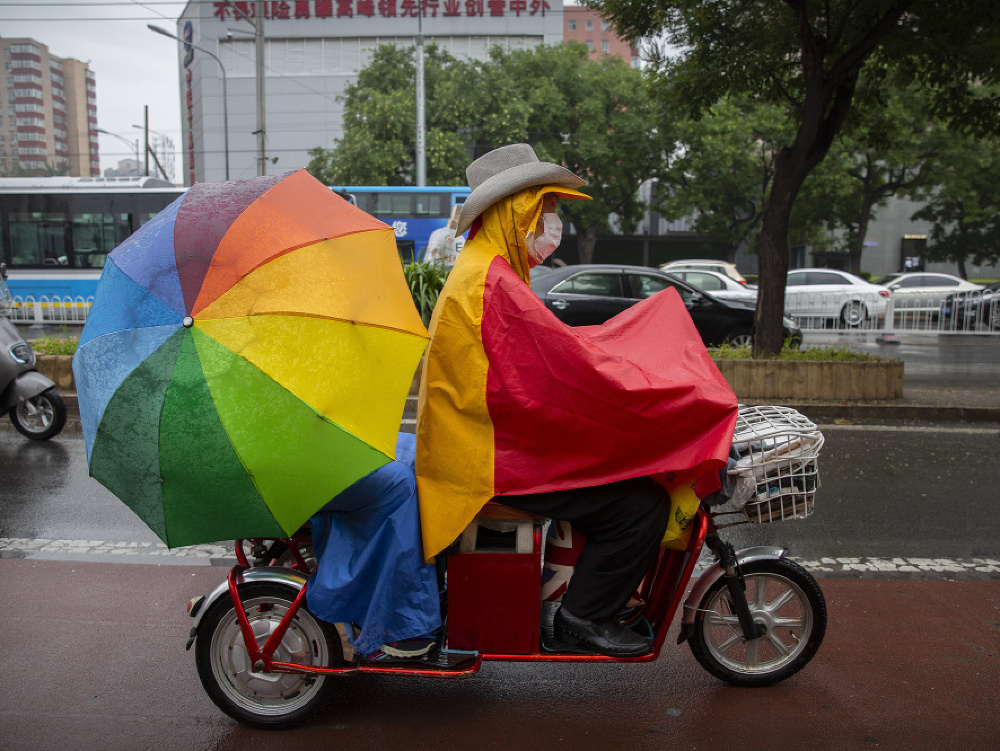 Muž s ochranným rúškom v pršiplášti ide na skútri v daždi v čínskom Pekingu.
