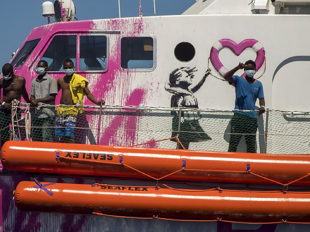 Chlapec s ochranným rúškom máva posádke záchranného plavidla z paluby ružovo-bielej motorovej jachty financovanej britským streetartovým umelcom Banksym.