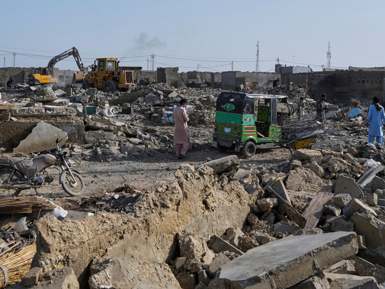 Na snímke ľudia prechádzajú cez trosky, zatiaľ čo hydraulická lopata búra budovu počas operácie proti nelegálnemu osídleniu afganských utečencov, ktorú vykonáva miestna vláda na okraji Karáčí v Pakistane.