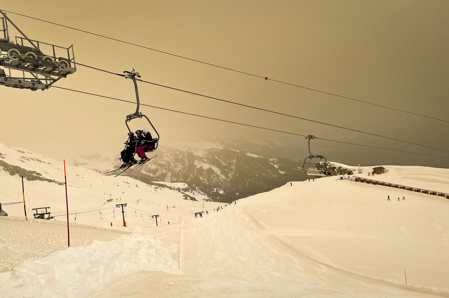 Lyžiari s ochrannými rúškami sa vezú lanovkou nad svahom pokrytým snehom, ktorý je sfarbený saharským pieskom na oranžovo a  vytvára špeciálnu svetelnú atmosféru vo švajčiarskom lyžiarskom stredisku Anzére