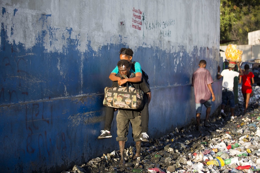 Muž s taškou v zuboch nesie ženu brodiac sa v odpadkoch a bahne po prietrži mračien v centre metropoly Port-au-Prince.