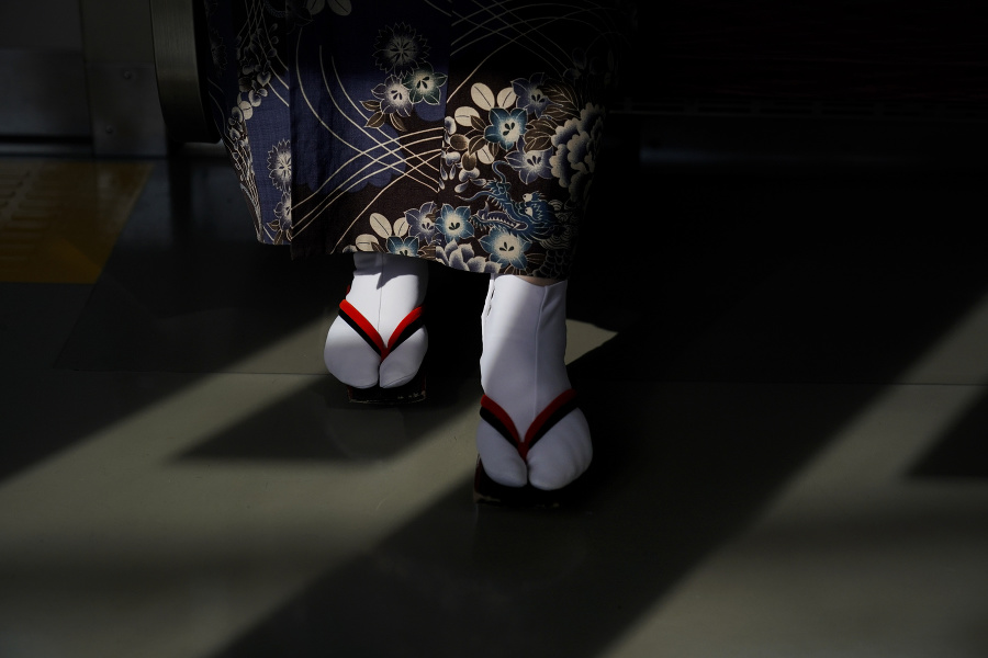 Tieň okna vo vlaku dopadá na ženu oblečenú v kimone a obutú v tradičných japonských sandáloch geta.