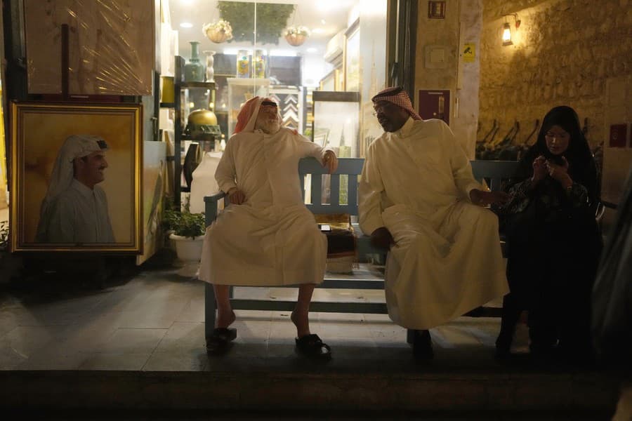 Portrét katarského emira, šejka Tamima bin Hamada Al Thaniho, je vystavený pred obchodom, kde dvaja muži a žena sedia na lavičke na trhu Souq Waqif.