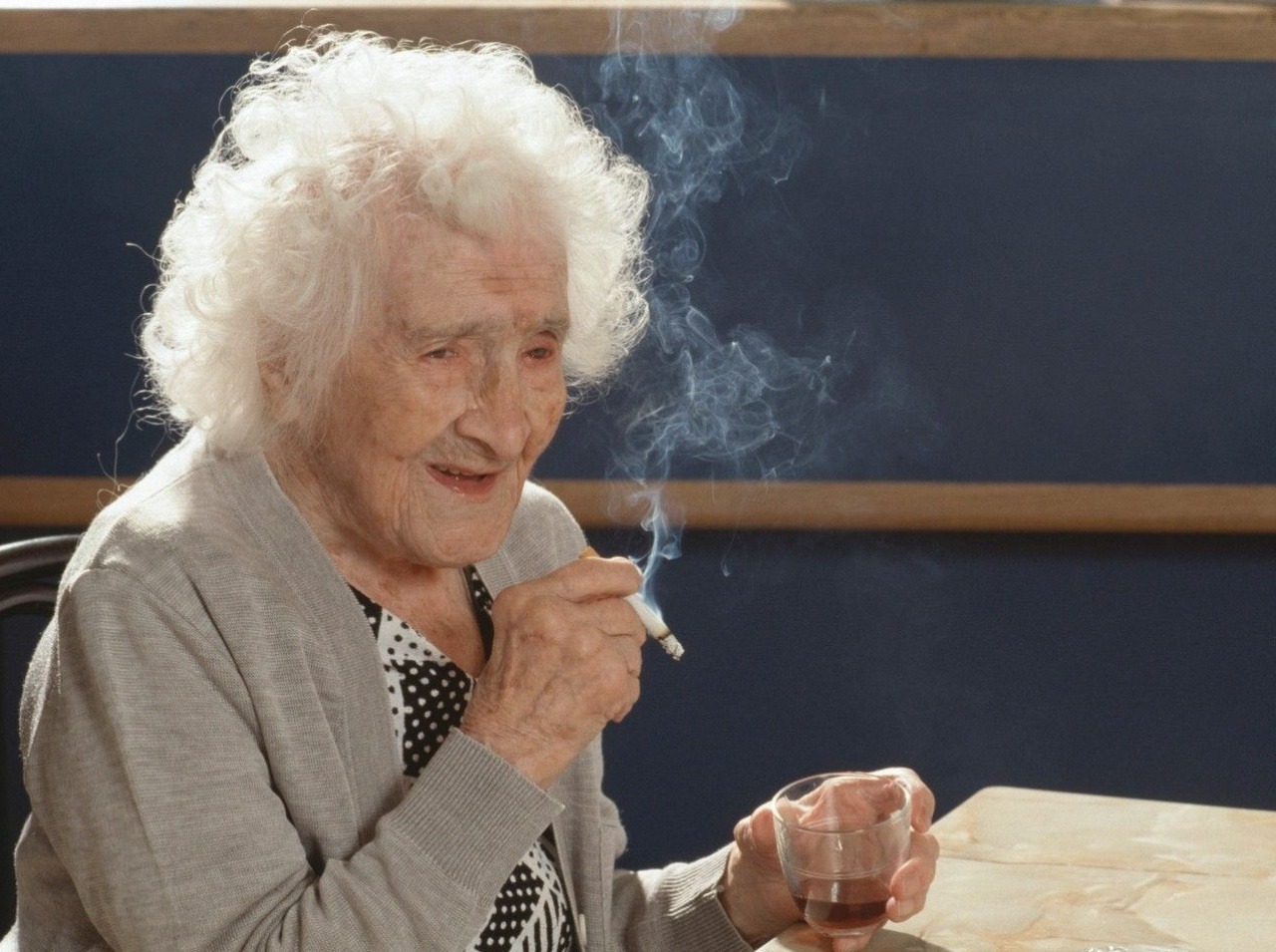 Jeanne Calmentová vo svojich 117-tich rokoch s cigaretkou, pohárikom alkoholu a úsmevom na tvári.